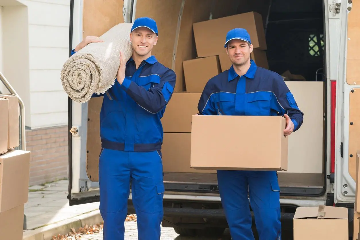 fm_f_jpex1G1 Two men in blue work clothes holding a roll of carpet and a box.