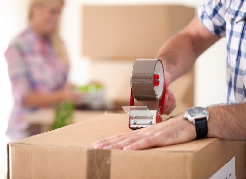 A person sealing a cardboard box with packing tape while another person is in the background.