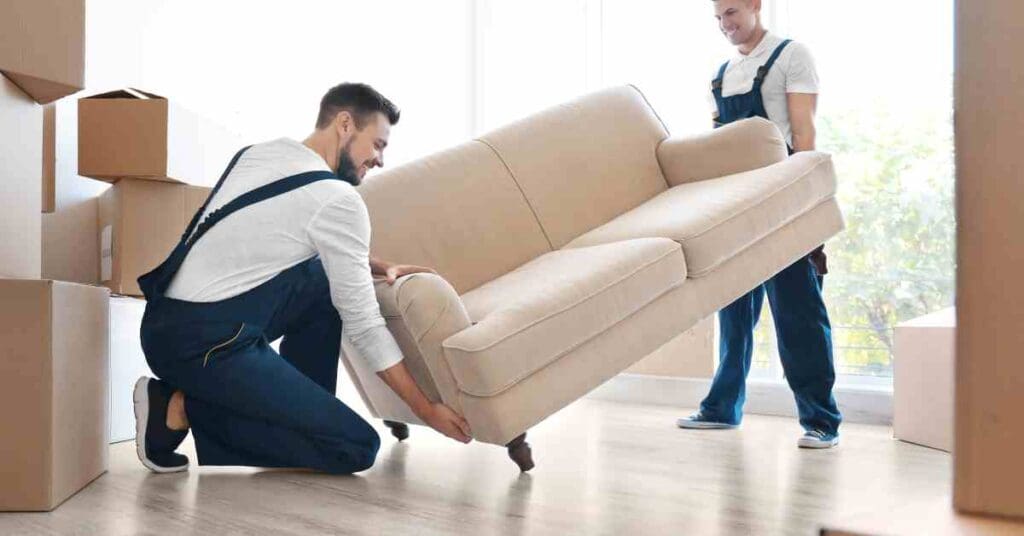 Two young men in white shirts and blue overalls are moving a large beige couch into a living room.