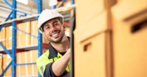 A friendly young man is in a storage warehouse full of boxes. He's smiling at the camera and pushing a trolley full of boxes.