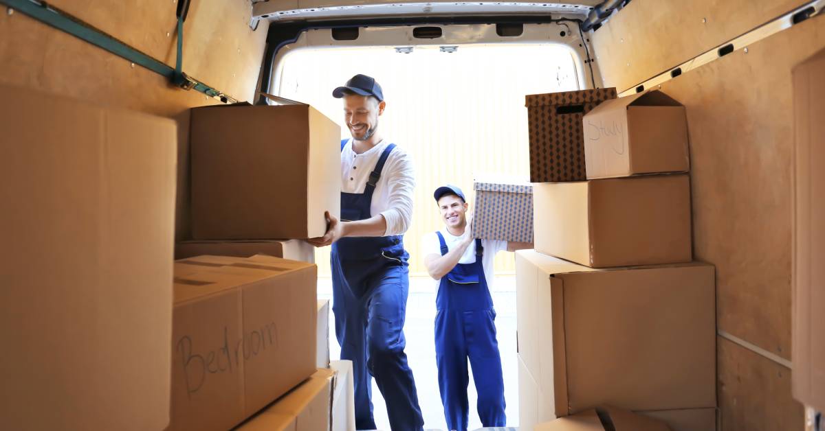 Two professional movers in overalls are climbing into the back of a moving van to unload it. The van is full of cardboard boxes.