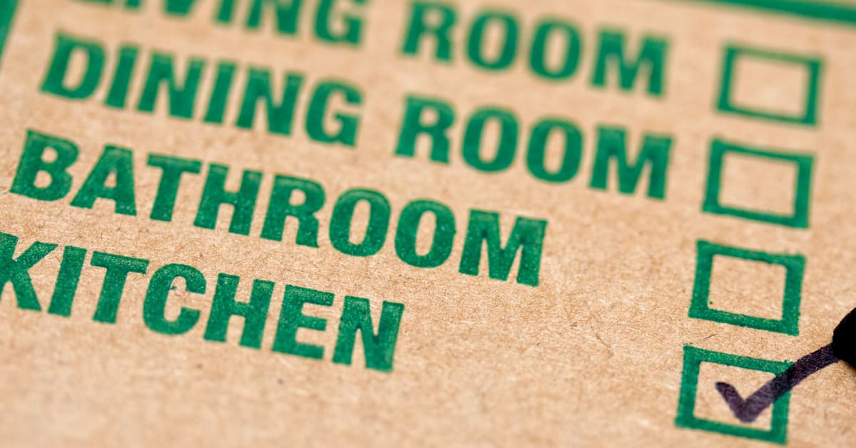 A cardboard moving box with text for different rooms: "Living Room. Dining Room. Bathroom. Kitchen" a checkmark is placed next to "Kitchen"