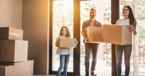 A family consisting of a mom, dad, and young daughter are moving into a new home. Each member of the family is carrying a cardboard box.