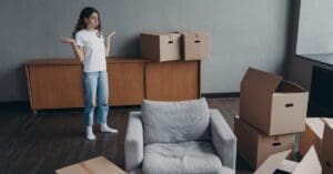 A woman standing in her living room surrounded by boxes and a gray chair. She's shrugging and exhaling while looking at the clutter.