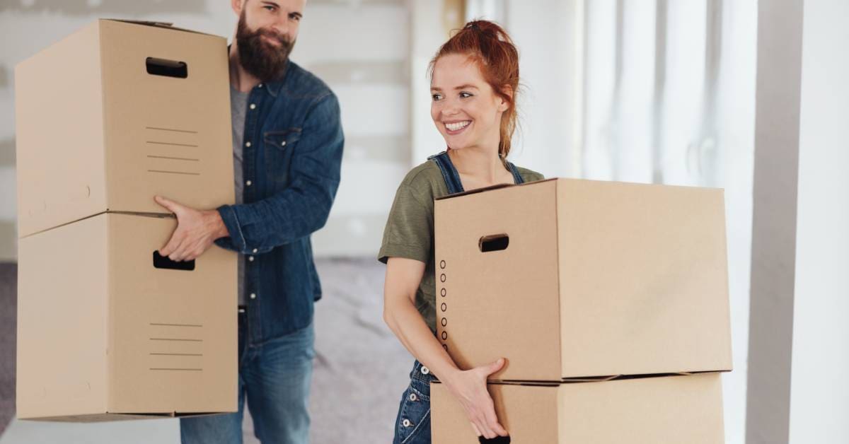 A young man and woman, both wearing denim, are each carrying a stack of two unlabeled brown cardboard boxes.