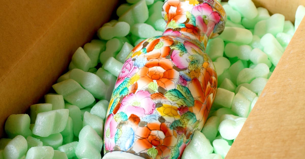 A floral vase with warm shades of pink, green, and yellow lying in a cardboard box of Styrofoam packing peanuts.