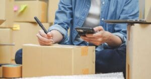 A person sits with legs crossed on the floor amid cardboard boxes, writing on one while holding a phone.