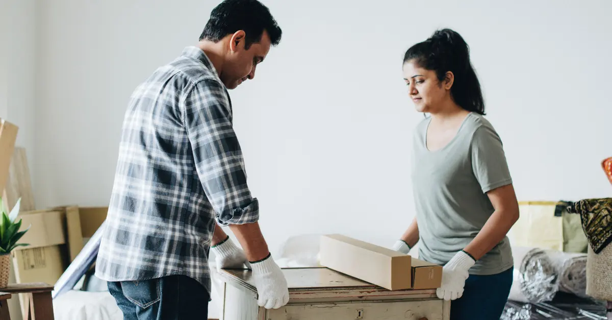 A man and woman wearing white gloves are lifting and carrying a white dresser with faded and cracked paint.