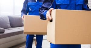 Two men in blue jumpsuits are carrying cardboard boxes in both hands. Behind them is a large gray couch.