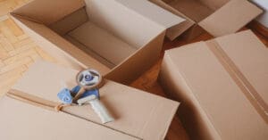Four cardboard boxes on a wooden floor. One is fully taped up while another has a tape gun resting on top of it.