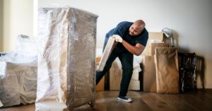 A man uses a roll of plastic wrap to wrap up a piece of furniture in a room filled with packaged items.