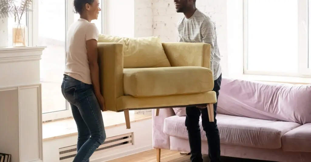 A young couple lifting and carrying a small yellow loveseat through their living room. A gray sofa sits in the background.