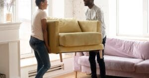 A young couple lifting and carrying a small yellow loveseat through their living room. A gray sofa sits in the background.