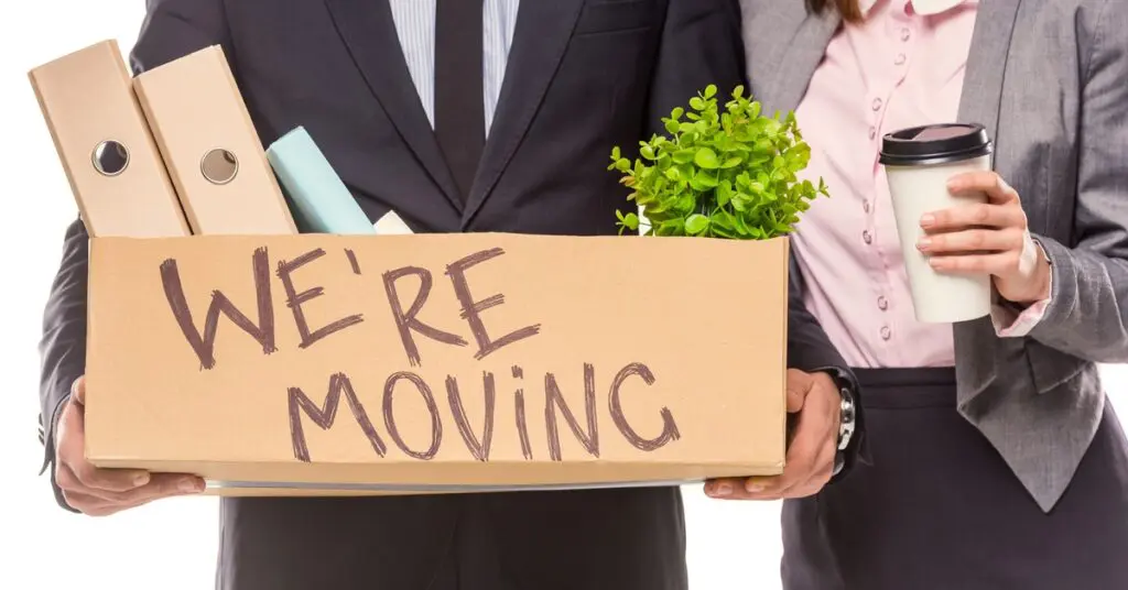 Two people wearing business attire. One is holding a cardboard box that says "We're Moving!" Binders and a plant are sticking out.