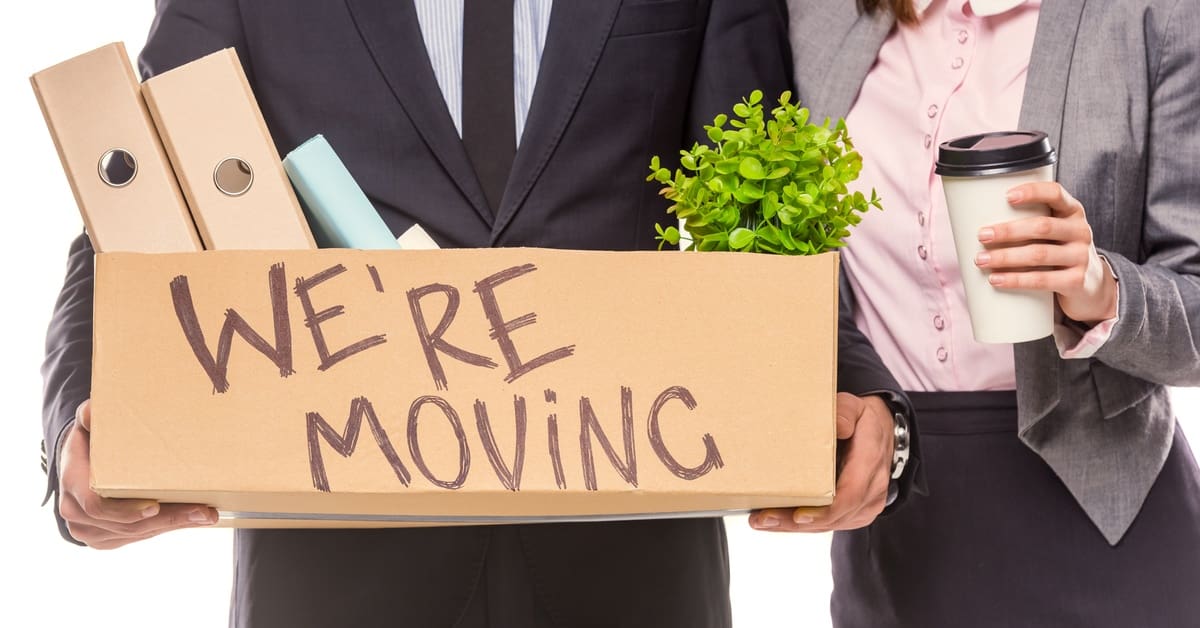Two people wearing business attire. One is holding a cardboard box that says "We're Moving!" Binders and a plant are sticking out.