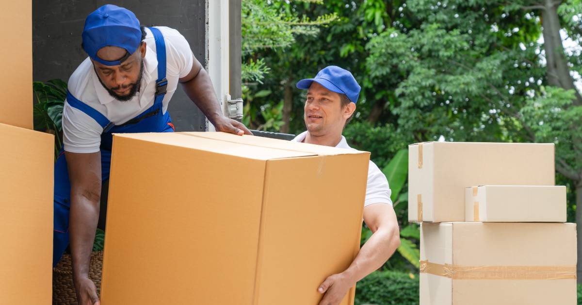 Two men in blue hats and overalls loading cardboard boxes onto the back of a large truck. The sun shines down on them.