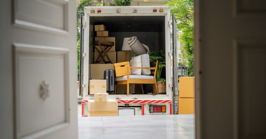A moving truck with the back open, as seen through a home's open front door. Inside the truck are boxes, chairs, and a rug.