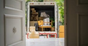A moving truck with the back open, as seen through a home's open front door. Inside the truck are boxes, chairs, and a rug.