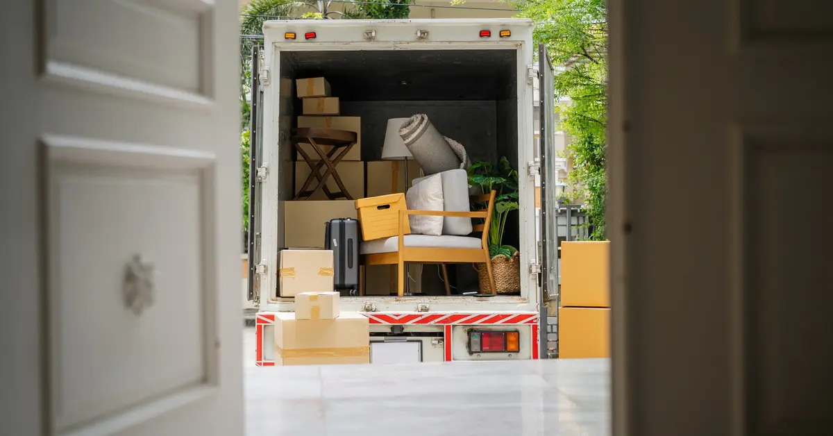 A moving truck with the back open, as seen through a home's open front door. Inside the truck are boxes, chairs, and a rug.