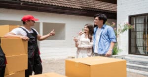 A man and woman are talking to a man in overalls next to a bunch of moving boxes. The man in overalls gives them a thumbs up.