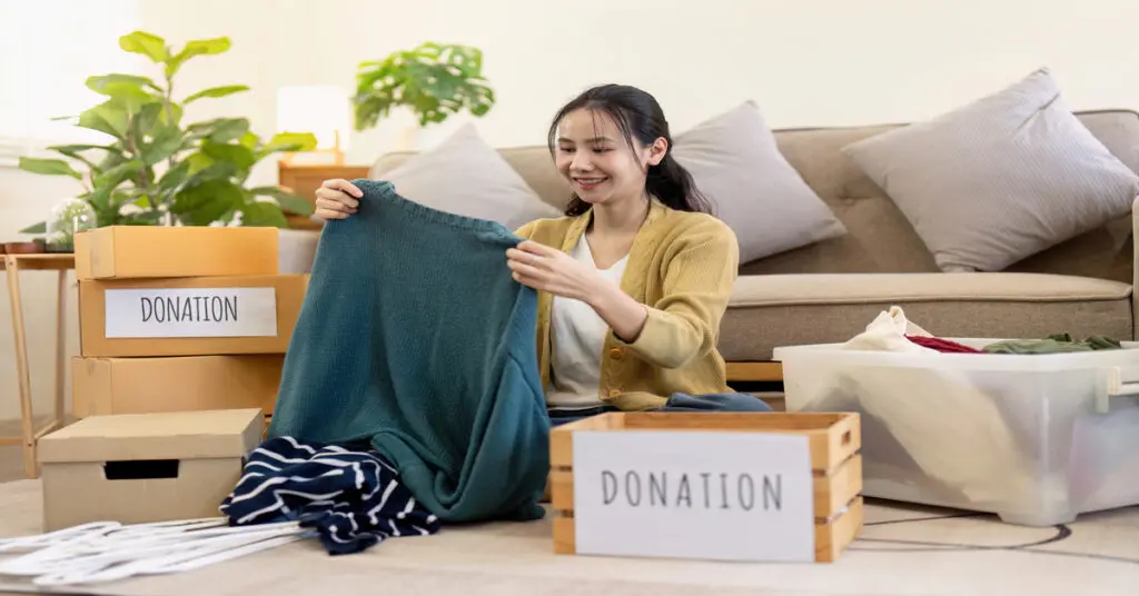 A woman is sitting on the floor of her living room and looking at a blue sweater. Next to her is a box labeled "Donation."