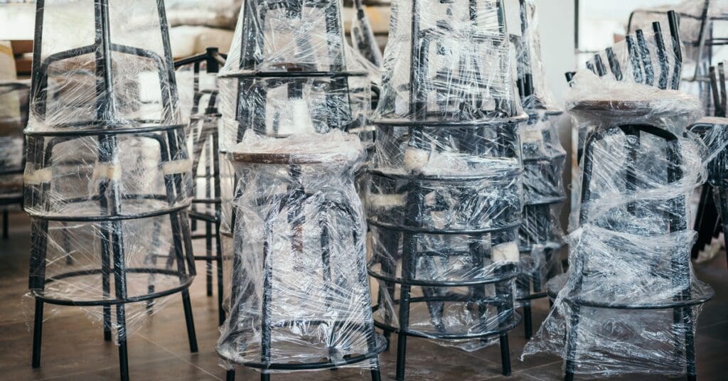 Four stacks of multiple black stools wrapped in tight plastic. The legs have extra cardboard padding around them.