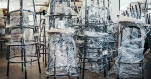 Four stacks of multiple black stools wrapped in tight plastic. The legs have extra cardboard padding around them.