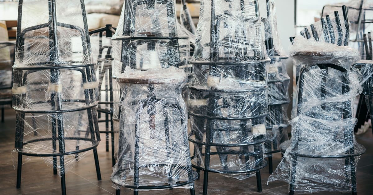 Four stacks of multiple black stools wrapped in tight plastic. The legs have extra cardboard padding around them.