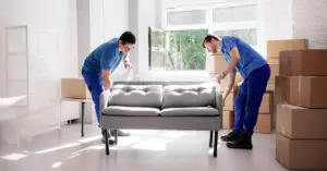 Two young men in similar blue clothes are lifting a gray couch in a living room. Off the side are several cardboard boxes.