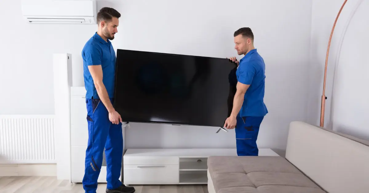 Two movers in blue jumpsuits hold a TV from both ends and lower it onto a white console in a sparsely furnished living room.