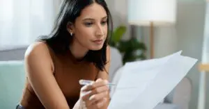 A woman sitting on a couch and leaning forward to examine a contract in one hand with a pen in the other.