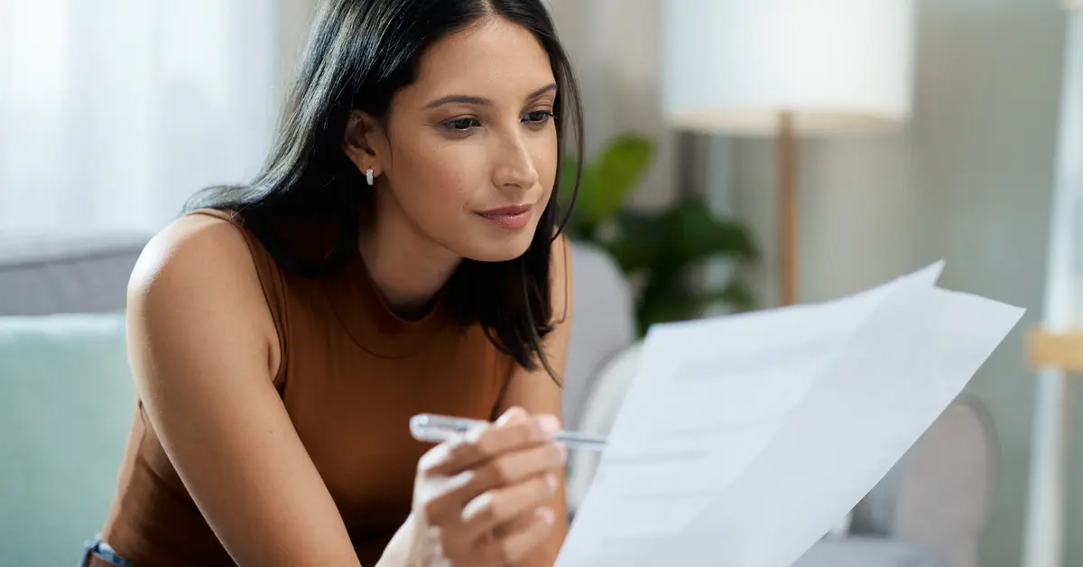 A woman sitting on a couch and leaning forward to examine a contract in one hand with a pen in the other.
