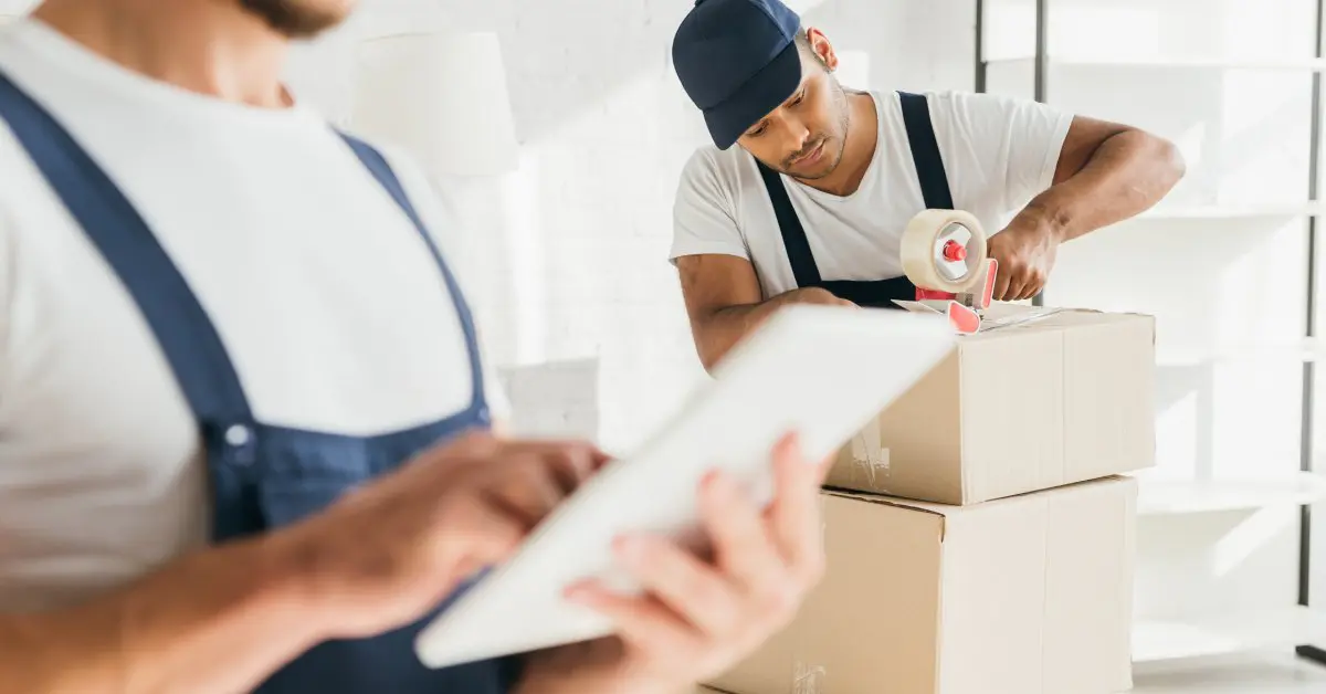 Two movers wearing white shirts and dark overalls are working. One is taping a box while the other is checking a clipboard.