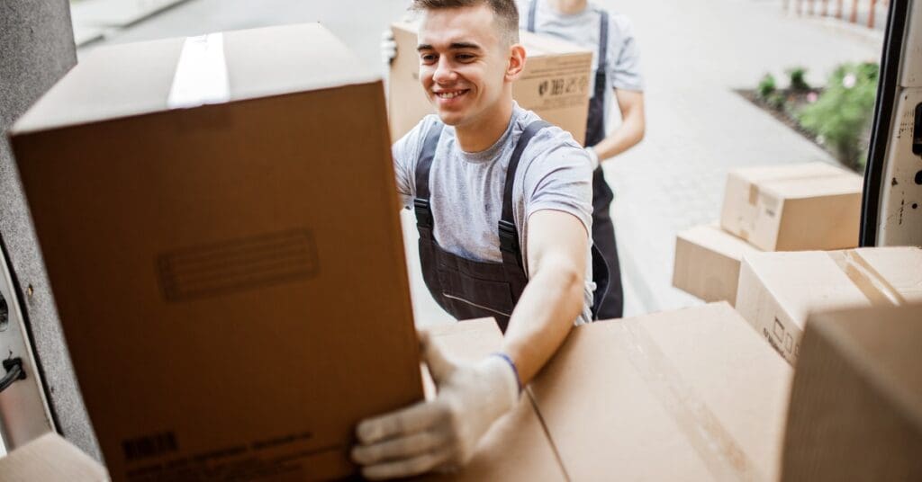 Two young professional movers in overalls carrying taped cardboard boxes and loading them into the back of a moving truck.