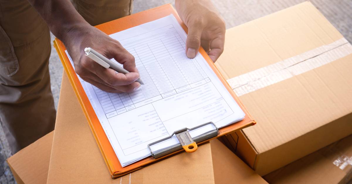 A close-up of a person writing on a data sheet on a clipboard, which rests on top of several taped cardboard boxes.