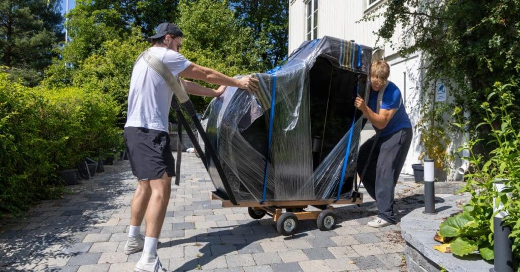 Two movers transporting a black grand piano. The piano is padded with rugs and shrink-wrap and sits on a dolly.